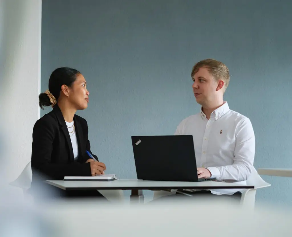 Eine Kollegin und ein Kollege von Köhler Transfer sitzen am Tisch mit Laptop und unterhalten sich.