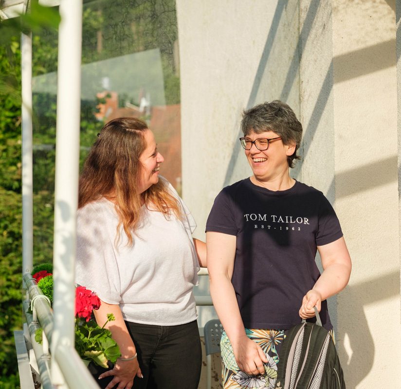 Zwei Frauen lachen gemeinsam auf einem sonnigen Balkon, umgeben von Blumen und Grün im Hintergrund.