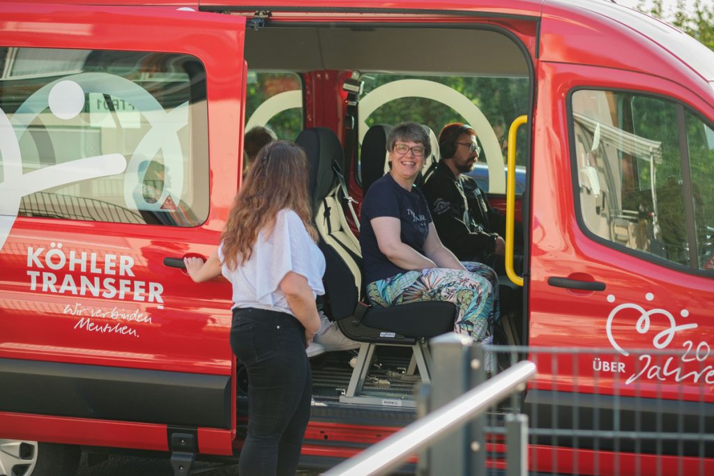 Eine Frau steigt fröhlich in ein rotes Köhler-Transfer-Fahrzeug, begleitet von einer freundlichen Begrüßung.