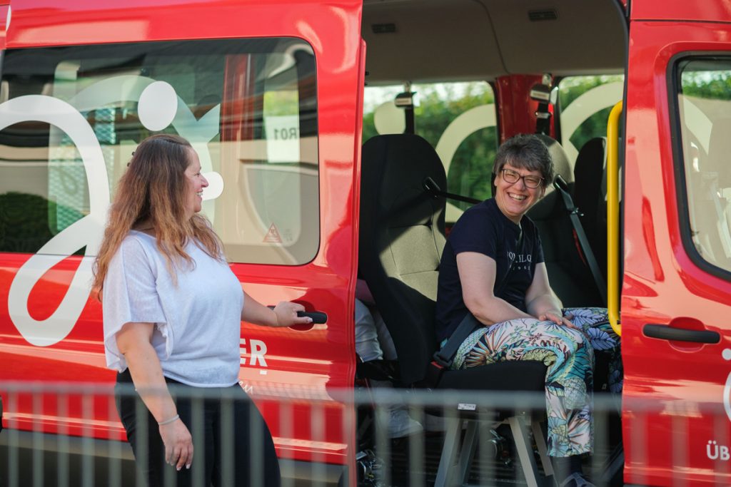 Zwei Frauen unterhalten sich lachend am roten Köhler-Transfer-Bus, die Stimmung ist gelöst.