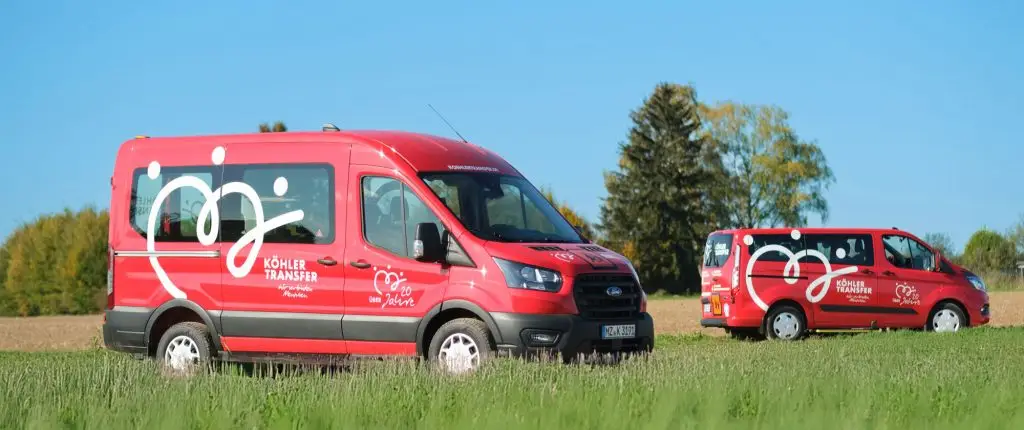 Zwei rote Köhler-Transfer-Busse stehen auf einer grünen Wiese, dahinter Bäume und blauer Himmel.