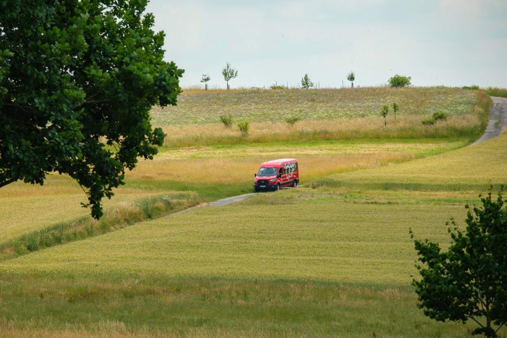 Ein roter Köhler-Transfer-Bus fährt allein über einen schmalen Weg durch eine grüne Feldlandschaft.
