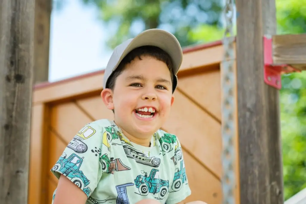 Ein lachender Junge mit grauer Kappe sitzt auf einem Spielplatz und trägt ein buntes T-Shirt mit Fahrzeugmotiven.