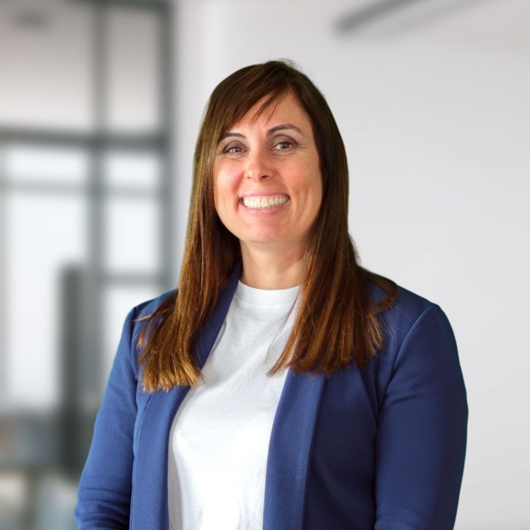 Frau mit langen braunen Haaren lächelt in einem hellen Büro, trägt blaues Sakko und weißes Shirt.